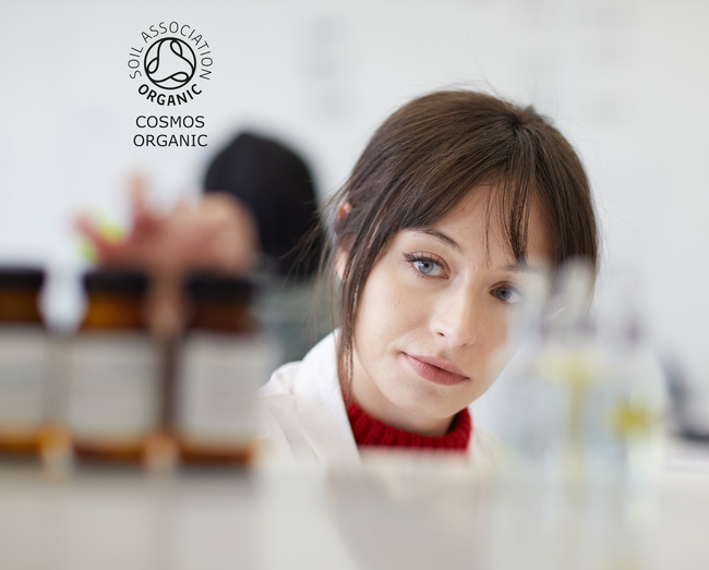 Image shows a shelf with products on being tested by a lab technician and the soil association organic Cosmos logo top left.